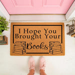 Book lover doormat with I Hope You Brought Your Books text and book stack illustrations, a natural coir welcome mat for front porch decor that makes a fun housewarming gift for readers and library fans on a white floor.