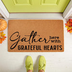 Fall welcome doormat featuring Gather Here With Grateful Hearts text on a natural coir fiber mat, perfect for autumn porch decor with colorful leaves and a pumpkin on a white floor in front of a bright green door for seasonal