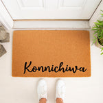 Konnichiwa coir doormat for front porch entry, featuring a natural fiber welcome mat with black script text, placed on a white floor with white sneakers visible, perfect for a stylish home entrance or housewarming decor.