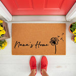 Nana's Home coir doormat featuring a black dandelion design, placed on a white porch in front of a red door.