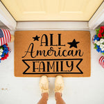 All American Family doormat featuring black text and stars on a natural coir fiber welcome mat, displayed on a front porch with patriotic red white and blue floral decor and a person standing in light-colored sneakers on the
