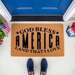 Patriotic God Bless America doormat for front porch entry, featuring natural coir fiber material with black text, perfect for a summer holiday welcome mat or outdoor home decor in front of a blue door with a small flag and