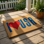 Patriotic USA doormat featuring a bold red heart graphic and blue lettering on a natural coir fiber welcome mat, perfect for a front porch entrance or outdoor home decor to show national pride and a festive American style look.