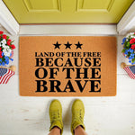 Patriotic coir doormat featuring Land of the Free Because of the Brave text and three stars, displayed on a front porch with flowers and flags, making a durable outdoor welcome mat for a home entrance or summer holiday decor.