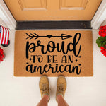 Proud to be an American doormat featuring black text and an arrow design on a natural coir fiber welcome mat, displayed on a front porch with patriotic decor for a festive entryway or housewarming entrance floor covering.