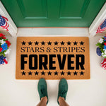 Patriotic Stars and Stripes Forever doormat featuring black text and stars on a natural coir fiber welcome mat, displayed on a front porch with festive red, white, and blue floral decor for a classic American home entryway look.