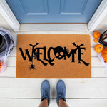 Spooky Halloween welcome doormat featuring a skull, bats, and spider web design on natural coir fiber, perfect for a festive front porch entry way decor during the fall season with pumpkins and cobwebs surrounding the entrance