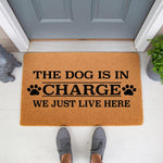 The Dog Is In Charge We Just Live Here doormat, a funny natural coir fiber welcome mat for dog lovers featuring black paw prints and bold text, placed on a front porch entryway for a humorous home decor look that greets every