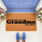 World's Best Grandma Lives Here coir doormat featuring bold black text on a natural fiber welcome mat, placed on a porch in front of a blue door with floral arrangements, perfect for a personalized home decor entry way floor rug.