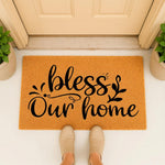 Bless Our Home doormat featuring black script text on a natural fiber coir rug, placed at a front door entrance with potted plants, creating a stylish welcome mat for housewarming decor and farmhouse porch entryways.