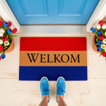 Welkom doormat with red, tan, and blue stripes, featuring a durable coir welcome mat design for a front porch entrance, styled with potted tulips and blue sneakers on a white floor, perfect for a colorful home entryway decor.