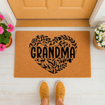 Grandma doormat featuring a black floral heart design on a natural coir welcome mat, placed on a porch floor with potted flowers and yellow sneakers, creating a charming entryway decor piece for a home entrance or front door.