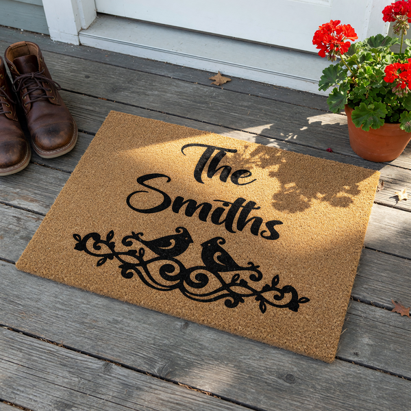 Personalized Family Name Coir Doormat featuring a custom last name in elegant black script, accented by a charming illustration of two birds perched atop a decorative scrollwork design on a natural tan fiber background.