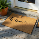 Welcome coir doormat featuring a natural tan fiber texture and elegant black script lettering that reads Welcome with a small heart icon, providing a warm and inviting front porch decor accent for any home entrance or entryway.