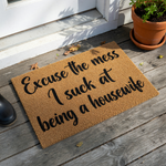 Funny Excuse the Mess I Suck at Being a Housewife doormat featuring bold black script lettering printed on a natural tan coir fiber mat, providing a humorous and welcoming touch to any front porch or entryway decor.