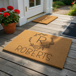 Personalized coir doormat featuring a custom last name and a large initial letter centered within a delicate black line art floral wreath, creating a welcoming entryway decor piece for a new home or a housewarming celebration.