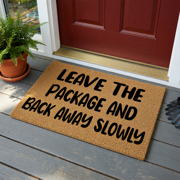 Funny Leave the Package and Back Away Slowly doormat featuring bold black text printed on a natural brown coir fiber mat, providing a humorous and functional welcome message for your front porch or entryway decor on a plain white