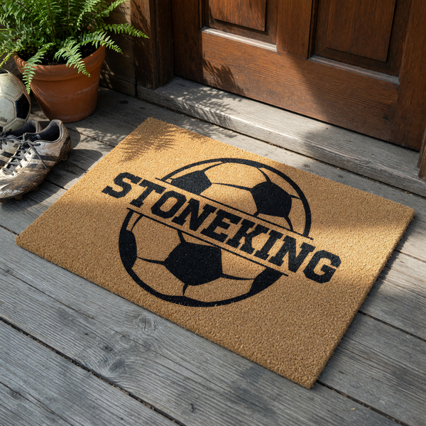 Personalized soccer ball doormat featuring a large black graphic of a soccer ball with a horizontal split across the center for a custom family name, set against a natural tan coir fiber background for a sporty outdoor entryway