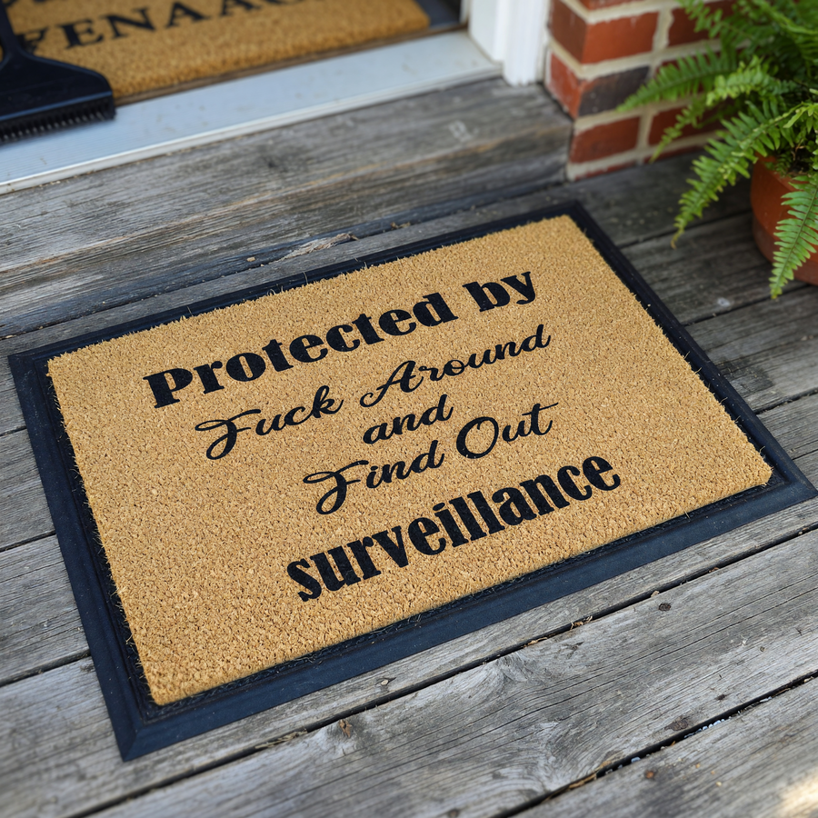 Funny doormat featuring the text Protected by Fuck Around and Find Out surveillance in a mix of bold and script fonts, displayed on a natural brown coir fiber mat against a clean white background for a humorous home entryway