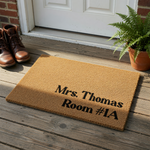 Personalized coir doormat featuring a natural tan fiber texture and bold black lettering that displays a custom name and room number, creating a functional and welcoming entrance accessory for a home, office, or classroom door.