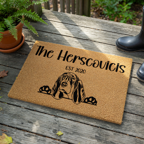 Personalized Basset Hound doormat featuring a natural coir texture with a black printed illustration of a Basset Hound peeking over the bottom edge, customized with a family name and established year on a clean white background.