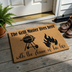 Grill Master coir doormat featuring a black graphic of a charcoal barbecue grill and a flame, with the text The Grill Master lives here with the flame of his life printed in a mix of bold and script fonts on a natural tan fiber